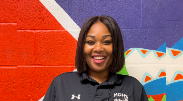 Middle-aged black women with a bob smiling, she is wearing a MOHS branded shirt and standing in front of a colorful brick wall